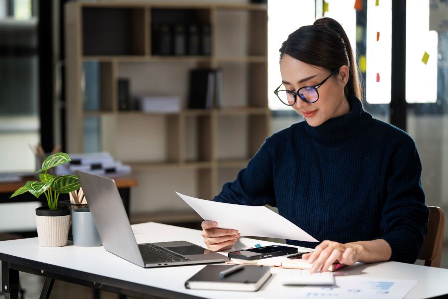 Woman working on laptop while reading paper. 