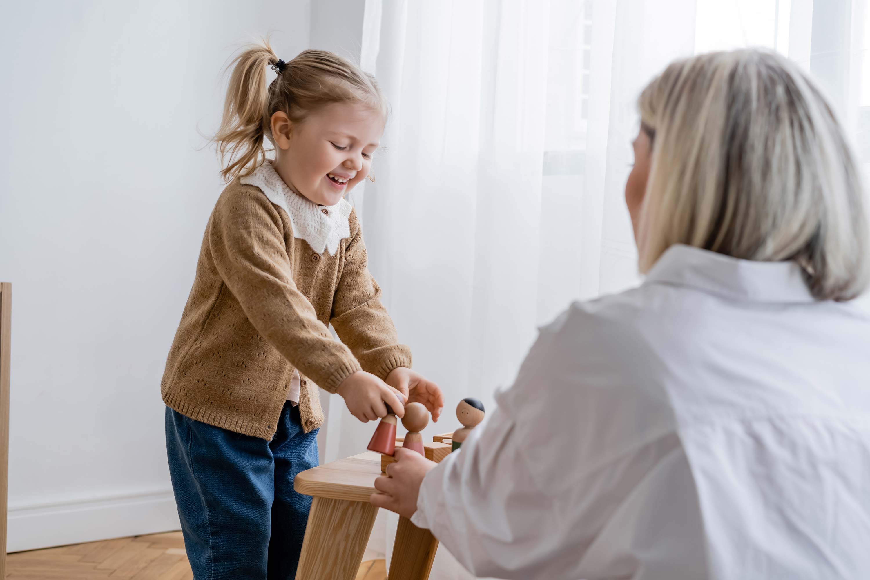 laughing child playing with wooden figurines near blurred mom at home