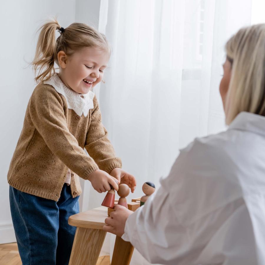 laughing child playing with wooden figurines near blurred mom at home